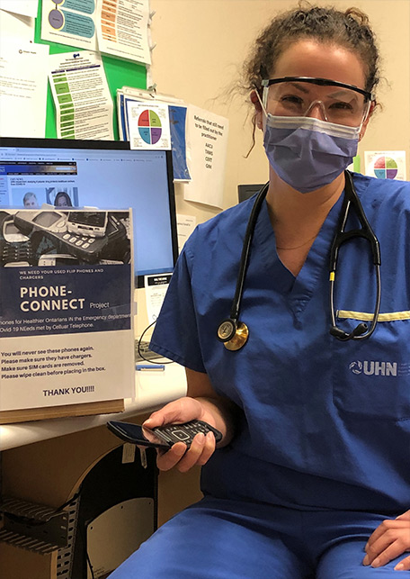 Dr. Andrea Somers in hospital scrubs, face mask and goggles. She is holding a phone, sitting at a desk and is smiling. Beside her is a small sign with information about the "Phone Connect" program.