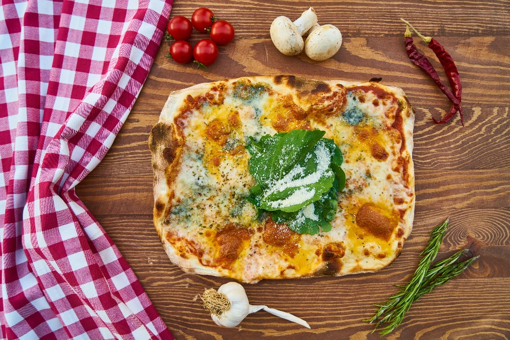 flat lay photo of a rectangle shaped pizza with various vegetables on a wooden table around it, on the left of the pizza there is a red and white checkered table cloth