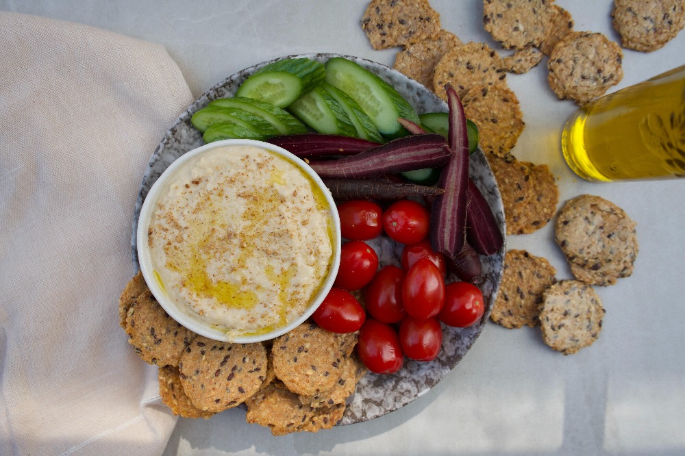 Flat lay photo of a plate with vegetables and a bowl of hummus. The plate is on a marble countertop, there is a beige linen cloth on the left hand side of the plate, on the right hand side of the plate there are extra crackers spilled across the table, there is also a bottle of olive oil on the right in partial view.